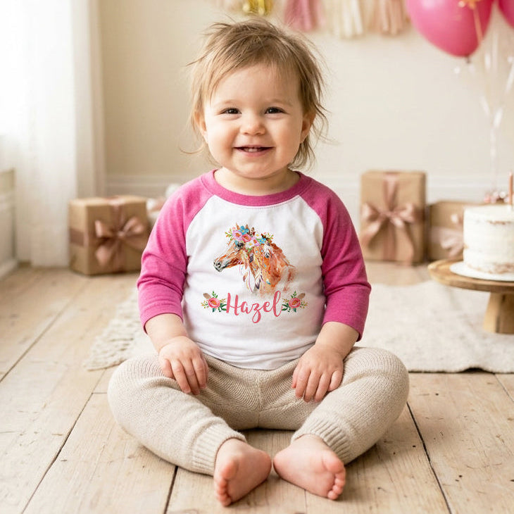 Child wearing a pink and white shirt with a horse boho design, sitting on a wooden floor with balloons and a cake in the background.
