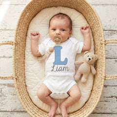Baby in a crib with a pacifier and teddy bear, wearing a onesie with 'Liam' on it.