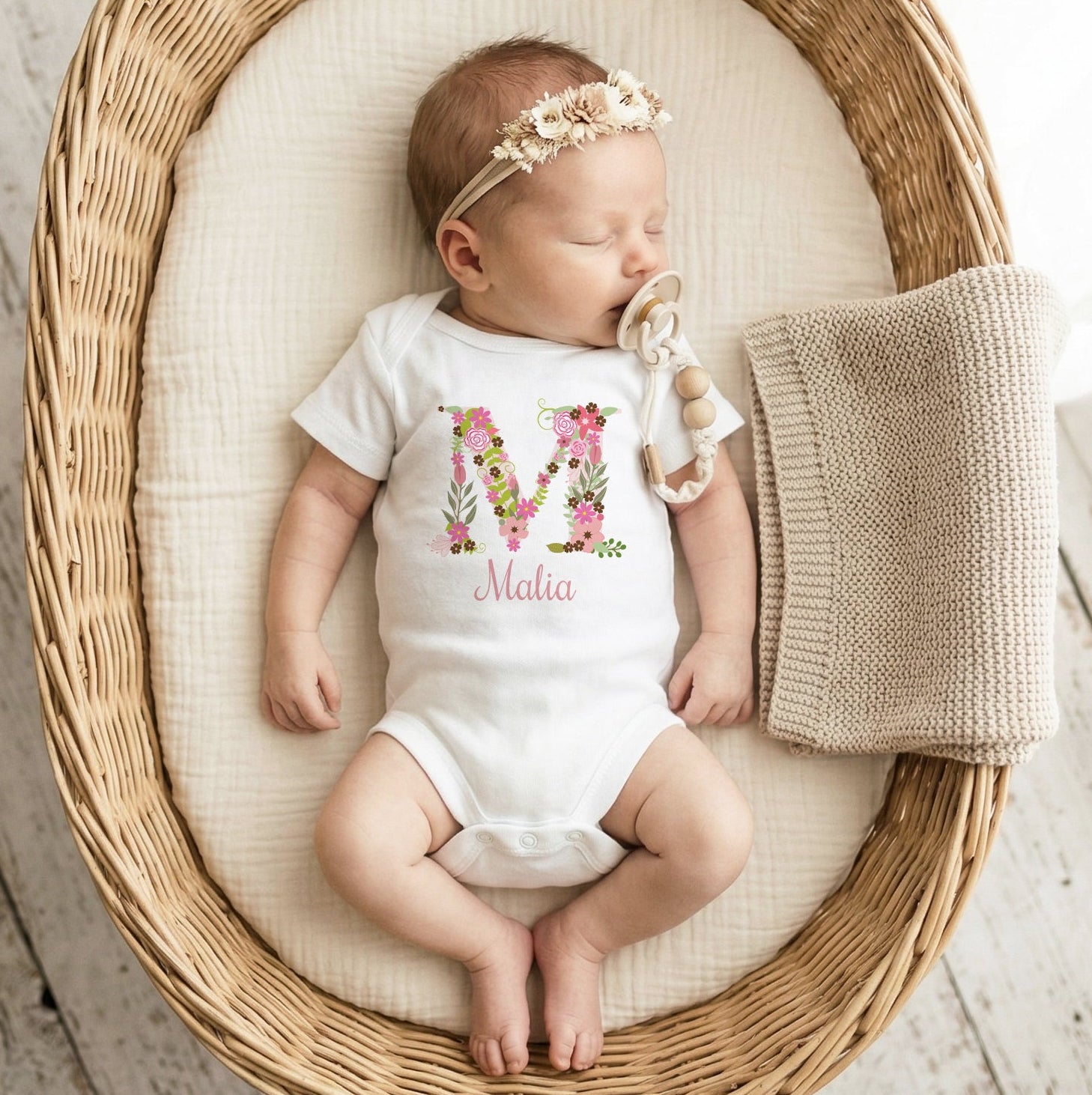 Baby in a white onesie with floral letter 'M' design, lying in a wicker crib on a wooden floor.