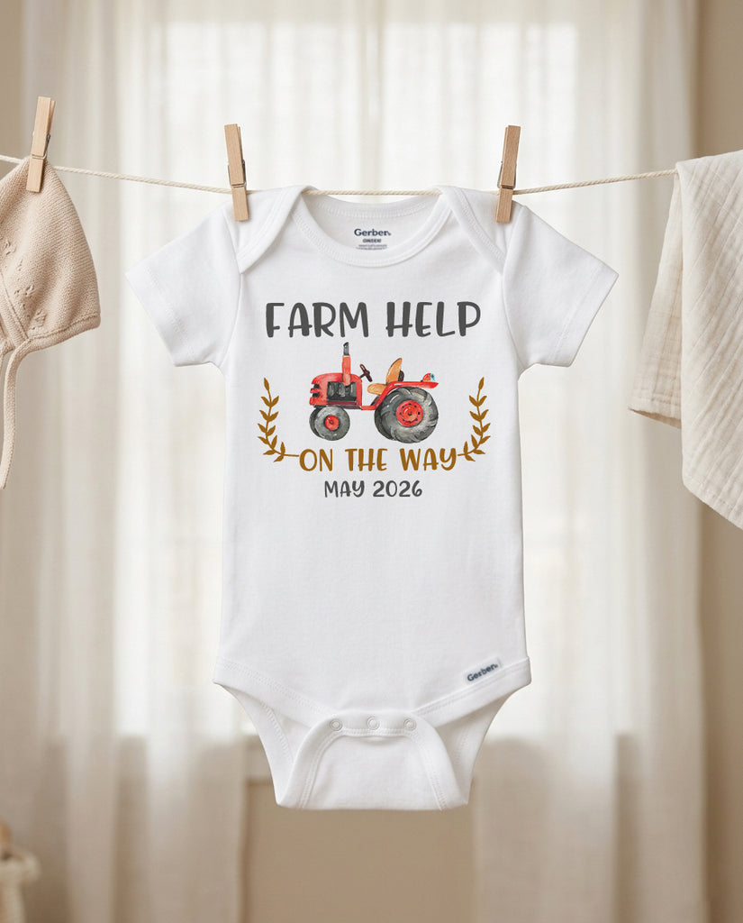 Baby onesie with 'Farm Help on the Way May 2026' text and tractor graphic hanging on a line.