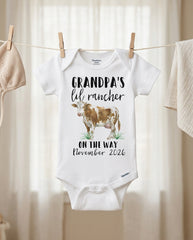 Baby onesie with 'Grandpa's lil' rancher on the way November 2026' text and cow illustration, hanging on a clothesline.