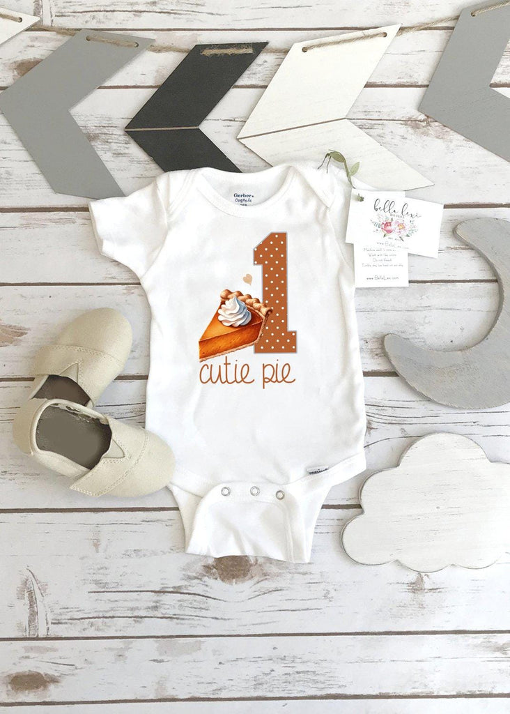 May include: A white baby onesie with a brown number one and a slice of pumpkin pie with whipped cream. The onesie has the words 'cutie pie' printed on it.