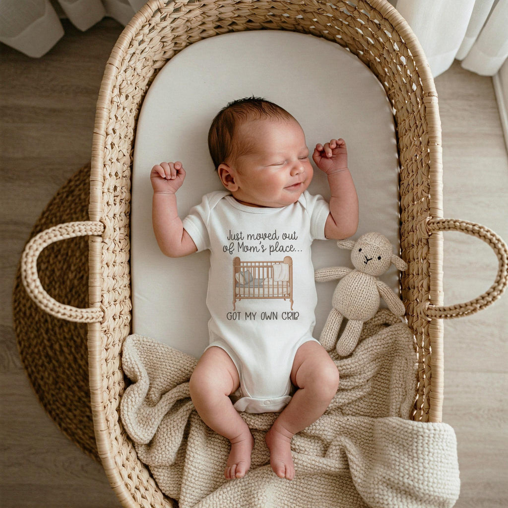 A newborn baby wearing a white onesie with the text 'Just moved out of Mom's place... GOT MY OWN CRIB' and a crib graphic. The baby is lying in a woven wicker bassinet with a cream-colored blanket. A small stuffed lamb toy is next to the baby.
