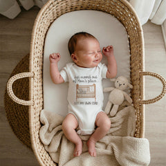 A newborn baby wearing a white onesie with the text 'Just moved out of Mom's place... GOT MY OWN CRIB' and a crib graphic. The baby is lying in a woven wicker bassinet with a cream-colored blanket. A small stuffed lamb toy is next to the baby.
