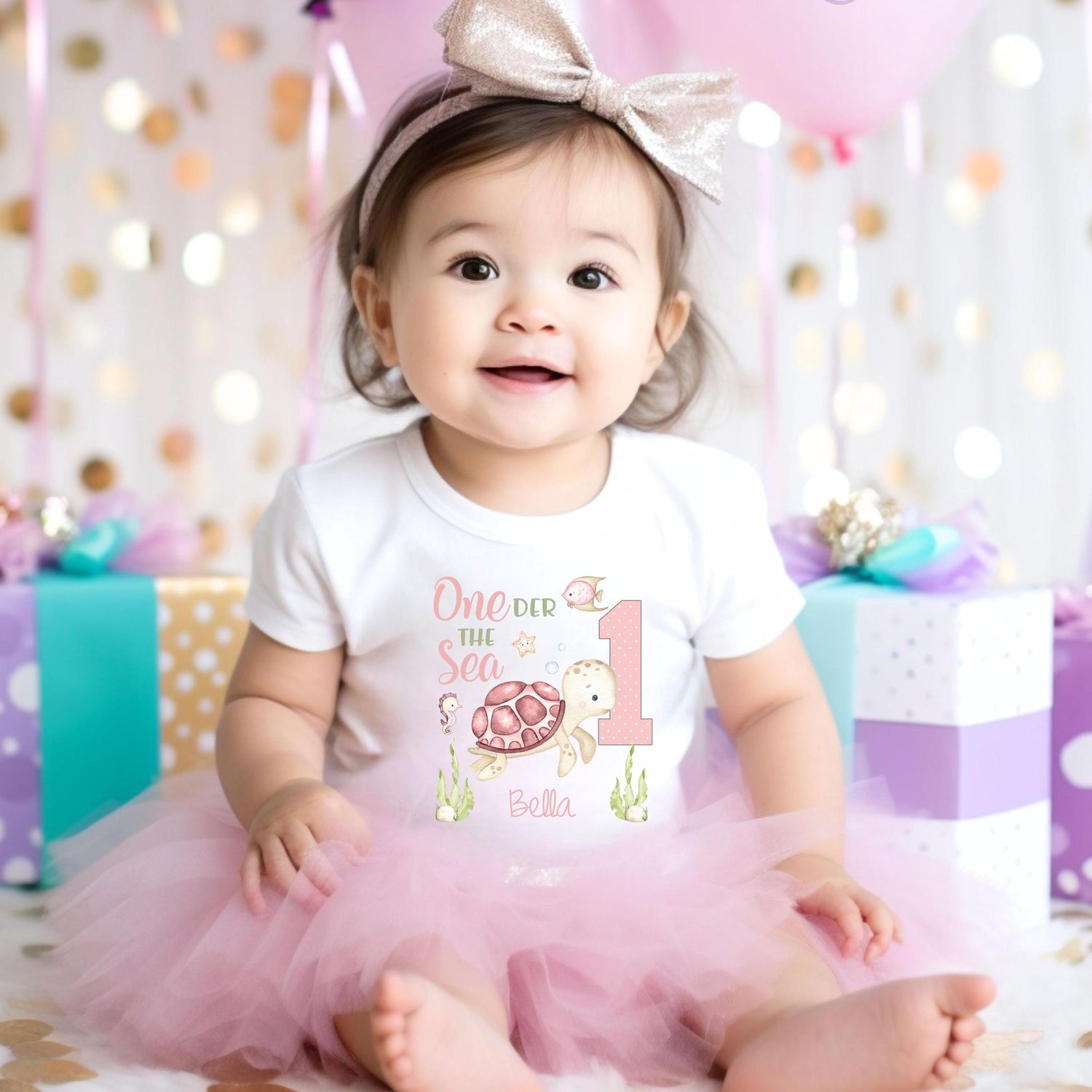 May include: A baby wearing a white onesie with a pink and green sea turtle design and the text 'One der the Sea' and the number '1'. The onesie also has the name 'Bella' printed on it. The baby is wearing a pink tulle tutu skirt.