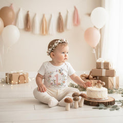 May include: A baby wearing a floral crown and a white onesie with a floral design and the number 1. The baby is sitting on a white floor with a small cake, mushroom decorations, and balloons. There are wrapped gifts in the background.