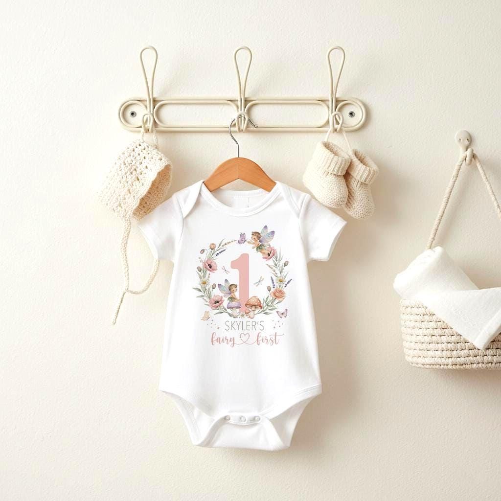 May include: White baby onesie with a floral wreath design, a pink number 1, and the text "Skyler's fairy first". The onesie is hanging on a wooden hanger, with a cream-colored hat and booties nearby.