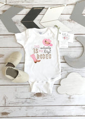 May include: White baby onesie with a pink cowboy hat and boots graphic. The text on the onesie reads "This is actually my first rodeo".