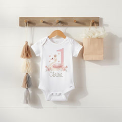 May include: A white short-sleeved bodysuit featuring a graphic of a swan wearing a crown, surrounded by pink flowers and butterflies. The number one is printed in a pink polka-dot pattern next to the swan, with the name Claire written below.