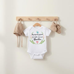 May include: A white short-sleeved baby bodysuit featuring a graphic of a blue bird, a pink heart, and a floral wreath with the text Un pequeño pajarito me contó, vas a ser Abuelita, accompanied by a matching beige knit bonnet and booties.