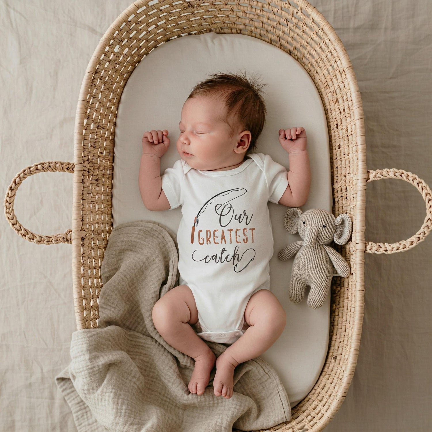 May include: A white short-sleeved baby bodysuit featuring a graphic of a fishing rod and the text Our GREATEST catch in black and brown ink. The bodysuit is worn by a sleeping infant lying in a woven wicker bassinet next to a small gray knit elephant.
