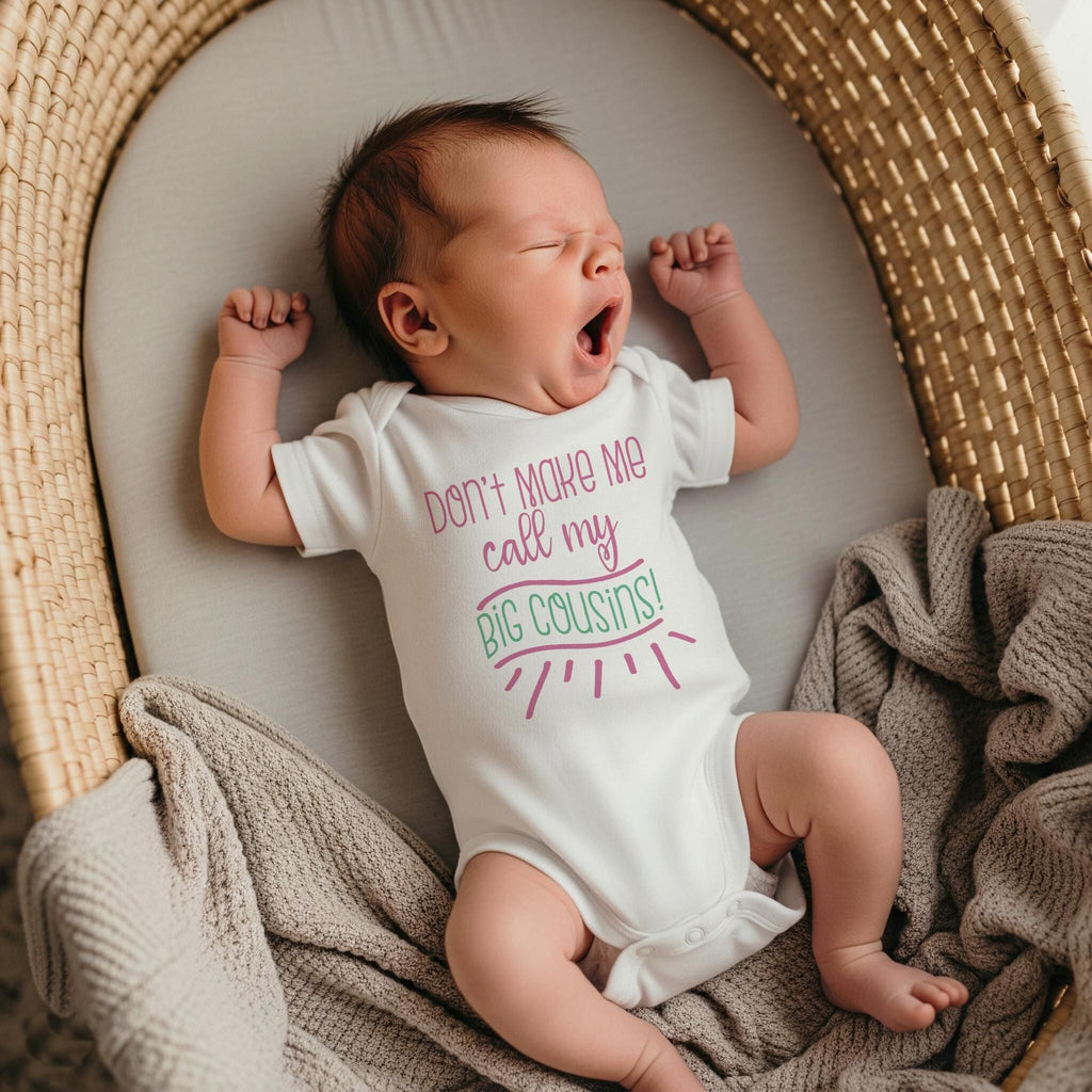 May include: A white short-sleeved baby bodysuit featuring the text Don't make me call my BIG COUSINS! printed in pink and green lettering on the chest. The garment has a round neckline and snap closures at the bottom for easy diaper changes.