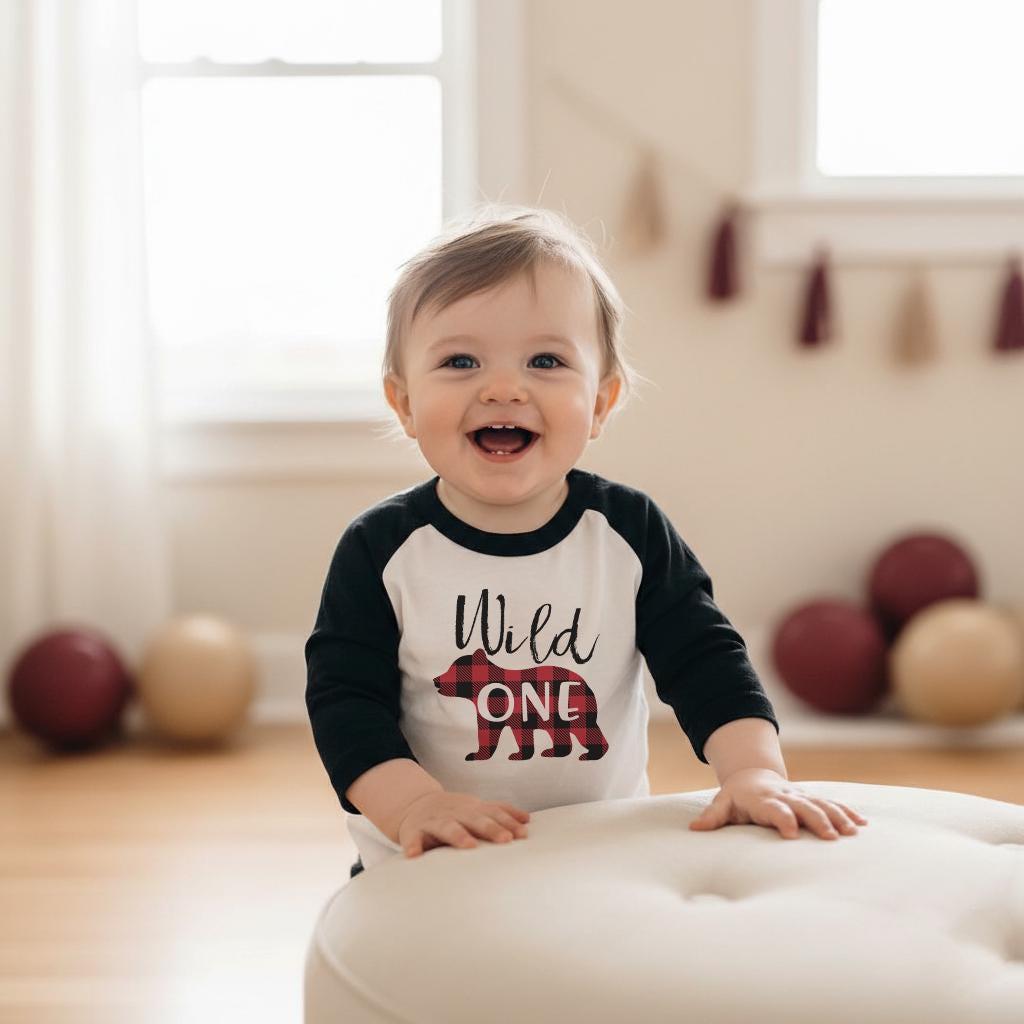 May include: A long-sleeve raglan shirt with black sleeves and a white torso. The front features the black text "Wild" above a graphic of a bear filled with a red and black buffalo plaid pattern, with the word "ONE" printed in white across the bear.