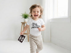 May include: A white short-sleeved bodysuit featuring the black text Big Brother est 2026 in a stylized font. The garment is worn with light beige pants. The wearer is holding a black and white ultrasound scan in one hand while smiling.