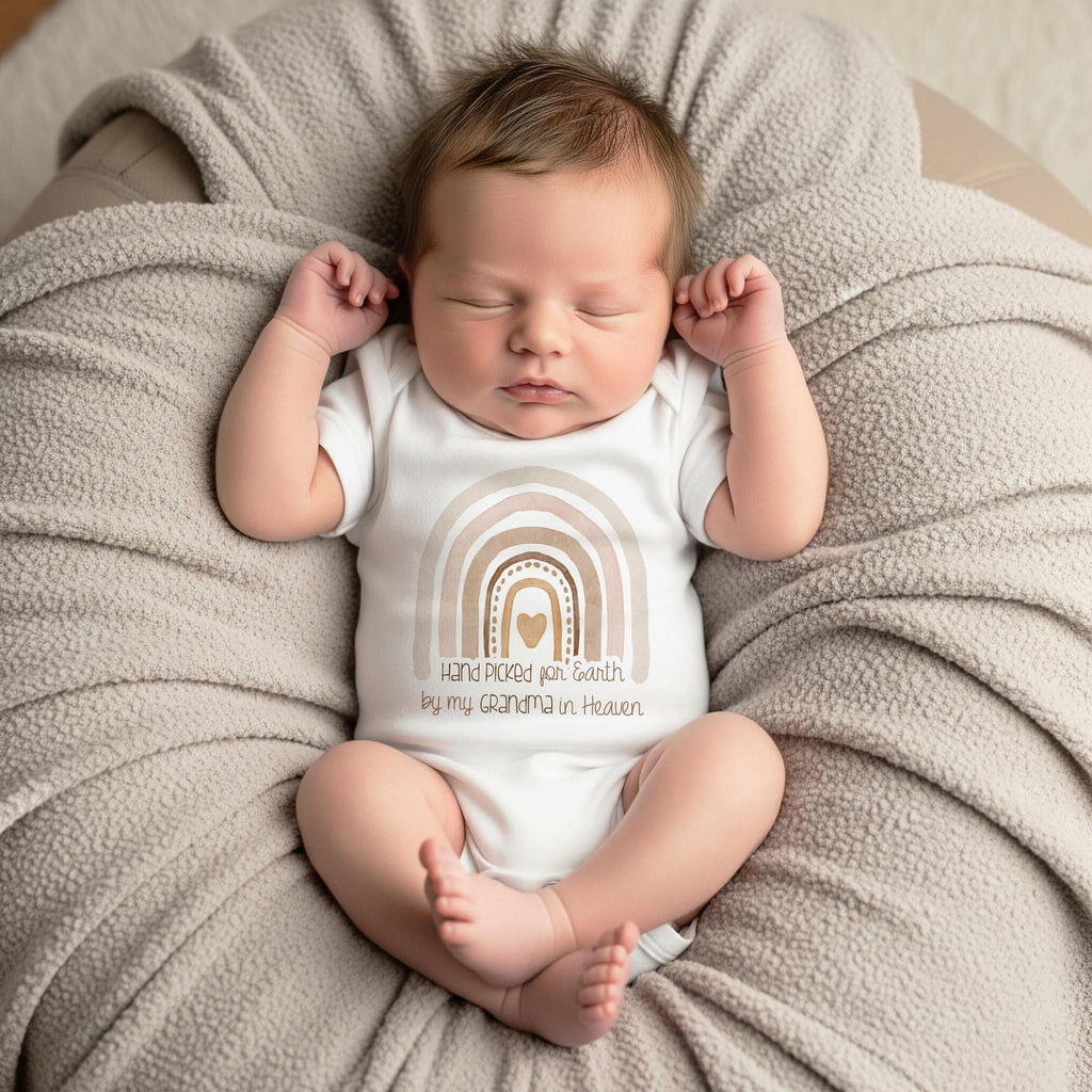 May include: A white short-sleeved baby bodysuit featuring a graphic of a multi-toned beige and brown rainbow with a central heart. Below the rainbow, the text reads: Hand Picked for Earth by my GRANDMA in Heaven, printed in a dark brown script.