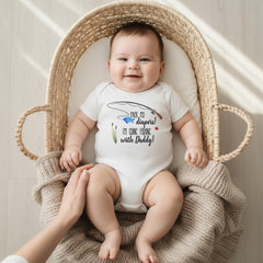 May include: A white short-sleeved baby bodysuit featuring a graphic of a fishing rod, a blue fish, and green reeds. The text reads: Pack my diapers! I'm going fishing with Daddy! The garment is made of soft fabric and has a round neckline.