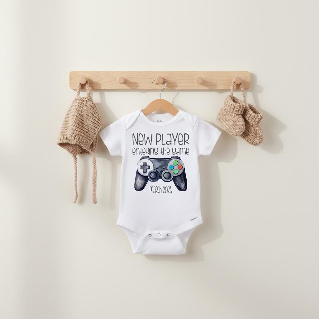May include: A white short-sleeved baby bodysuit featuring a black graphic of a video game controller and the text "NEW PLAYER ENTERING THE GAME MARCH 2026". The bodysuit is displayed on a wooden hanger with a matching beige knit bonnet and booties.