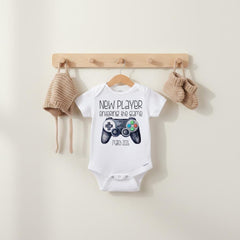 May include: A white short-sleeved baby bodysuit featuring a black graphic of a video game controller and the text "NEW PLAYER ENTERING THE GAME MARCH 2026". The bodysuit is displayed on a wooden hanger with a matching beige knit bonnet and booties.