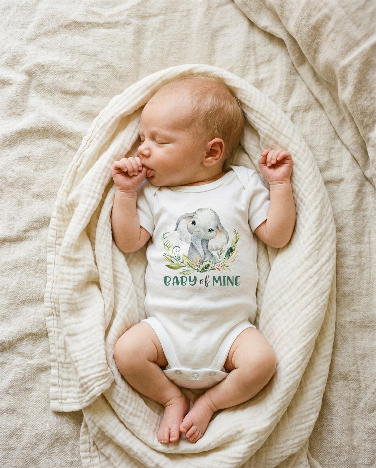 May include: A white short-sleeved baby bodysuit featuring a watercolor illustration of a gray elephant surrounded by green leaves and the text BABY OF MINE printed in green letters on the front, worn by a sleeping infant wrapped in a cream blanket.