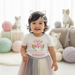 May include: A white short-sleeved t-shirt featuring a graphic of a cat face adorned with pink flowers and the text Two Purrfect! The shirt is paired with a multi-colored pastel tulle skirt and a thin gold-toned cat ear headband accessory.
