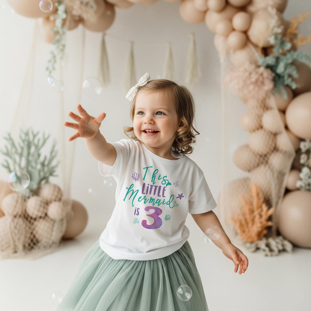 May include: A white short-sleeved t-shirt featuring the text This Little Mermaid is 3 in teal and purple lettering, accented with small purple and teal seashell and starfish graphics, paired with a sage green layered tulle skirt.