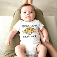 May include: A white short-sleeved baby bodysuit featuring a graphic of yellow nachos with green jalapeño slices and black olives. The text reads DO NOT kiss me if I'm NACHO BABY in black lettering centered on the chest of the garment.