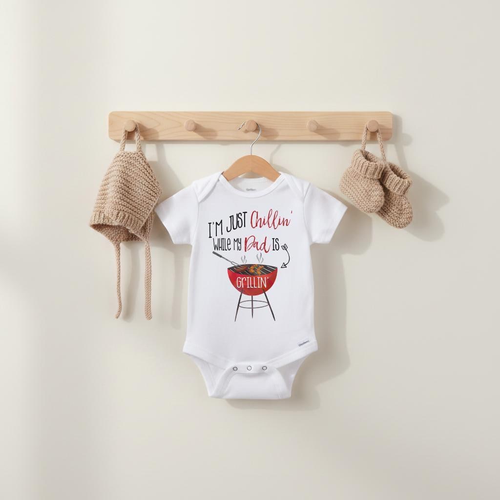 May include: A white short-sleeved baby bodysuit featuring the text I'M JUST Chillin' WHILE MY Dad IS GRILLIN' and a red charcoal grill graphic. The bodysuit is displayed with a matching tan knit bonnet and a pair of tan knit baby booties.