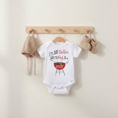 May include: A white short-sleeved baby bodysuit featuring the text I'M JUST Chillin' WHILE MY Dad IS GRILLIN' and a red charcoal grill graphic. The bodysuit is displayed with a matching tan knit bonnet and a pair of tan knit baby booties.