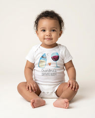 May include: A white short-sleeved baby bodysuit featuring a graphic of a baby bottle and a wine glass separated by a small orange heart. Below the graphic, the text reads Grandma's DRINKING BUDDY in a mix of script and block lettering.