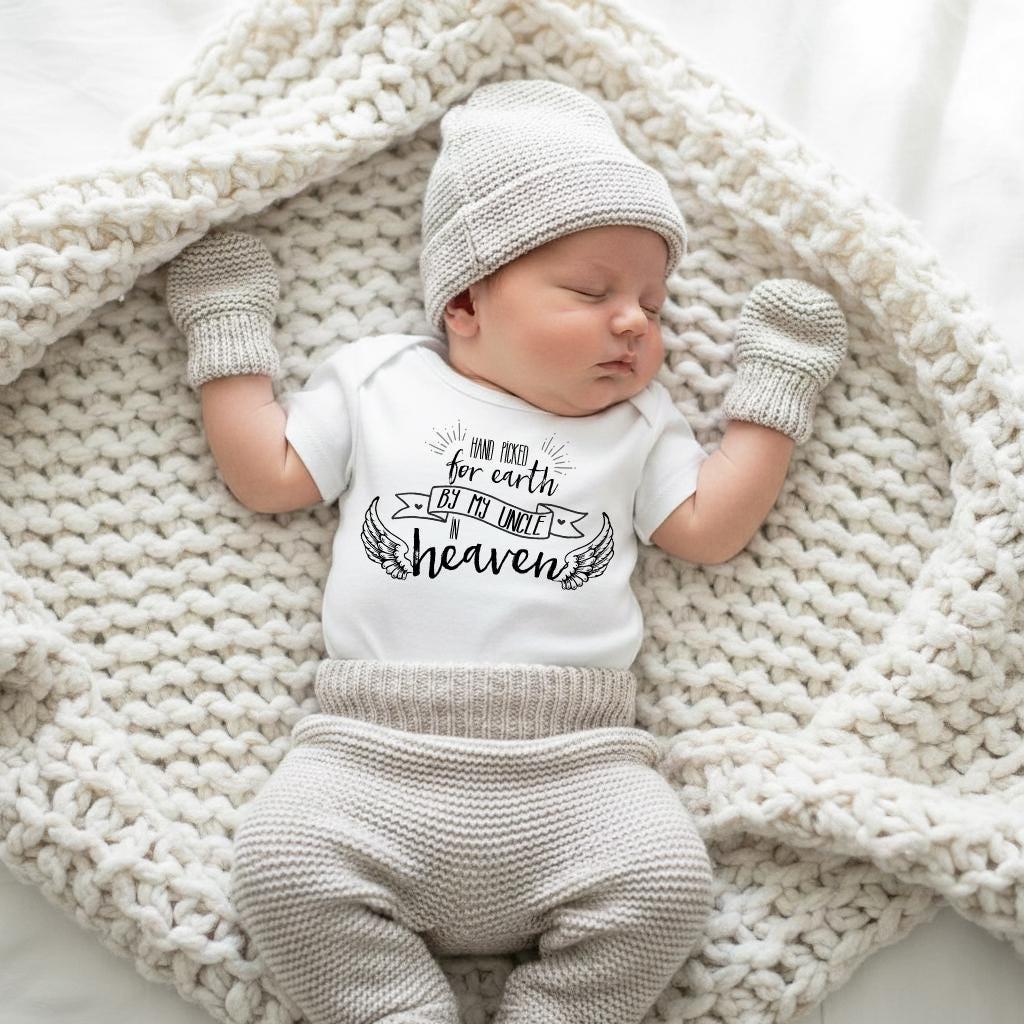 May include: A white short-sleeved bodysuit with black text that reads Hand picked for earth by my uncle in heaven, featuring wing illustrations. The outfit includes a matching light gray knit beanie, knit mittens, and knit pants.