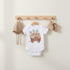 May include: White baby onesie with a colorful illustration of vegetables in a red wagon, with the text "Locally Grown". A beige knit hat and booties hang on a wooden rack. The onesie is hanging on a wooden hanger.