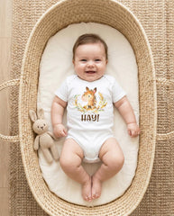 May include: A white short-sleeved baby bodysuit featuring a watercolor illustration of a brown horse surrounded by wheat stalks and the text HAY! printed in brown. A small beige crocheted rabbit toy is positioned next to the bodysuit.