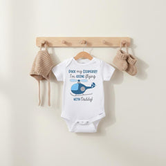 May include: A white short-sleeve baby bodysuit featuring a blue helicopter graphic and the text "PACK my DIAPERS!! I'm GOING flying WITH Daddy!". The bodysuit is displayed with a matching tan knit bonnet and a pair of tan knit baby booties.
