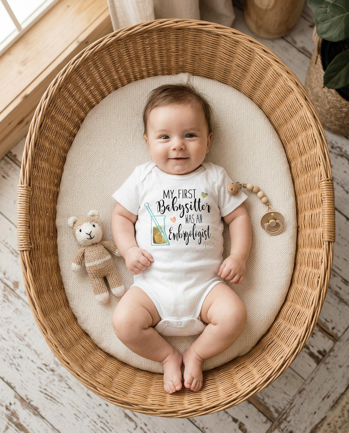 May include: A white short-sleeved baby bodysuit featuring the text 'MY FIRST Babysitter WAS AN Embryologist' in black script and print, accompanied by a graphic of a laboratory beaker with a gold liquid and a small gold heart icon.