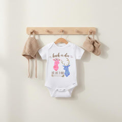 May include: A white short-sleeved baby bodysuit featuring a pink watercolor deer silhouette and a blue watercolor deer silhouette with the text "buck or doe" and "can't wait to know" above and below the deer, accompanied by a knit hat and booties.