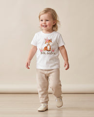 May include: A white short-sleeved t-shirt featuring a graphic of a fox adorned with pink flowers and the text BIG Sister in brown script, paired with beige knit trousers and light-colored shoes. The shirt has a crew neckline and a straight hem.