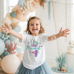 May include: A white short-sleeved t-shirt featuring the text This LITTLE Mermaid is 4 in teal and purple script. The design includes small illustrations of a seashell, a starfish, and a mermaid tail. The shirt is paired with a blue tulle skirt.