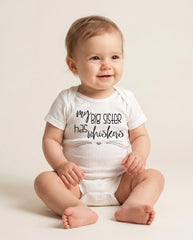 May include: A white short-sleeved baby bodysuit featuring the black text 'my BIG SISTER has whiskers' printed on the chest, accompanied by a small heart icon and thin horizontal lines resembling cat whiskers below the lettering.