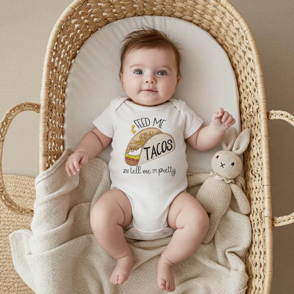 May include: A white short-sleeved baby bodysuit featuring a graphic illustration of a taco and the text "FEED ME TACOS and tell me I'm pretty" in black lettering. The garment is worn by an infant lying in a woven bassinet with a beige blanket.