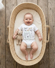 May include: A white short-sleeved baby bodysuit featuring the text Don't make me call my Auntie and a graphic of a smartphone. A small beige crocheted teddy bear is positioned next to the bodysuit, which is resting inside a woven basket.