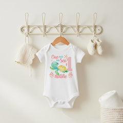 May include: A white short-sleeved baby bodysuit featuring a graphic of a green sea turtle, a starfish, and coral. The text reads One der the Sea 1 and the name Natalie in pink and teal script. A matching white crocheted bonnet and booties are included.