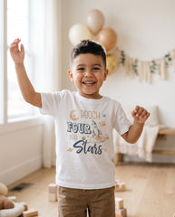 May include: A white short-sleeved t-shirt featuring a graphic print with a rocket, a smiling moon, planets, and the text "Reach FOUR THE Stars" in blue and tan colors. The design is centered on the chest of the garment.