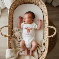 May include: A white short-sleeved baby bodysuit featuring a watercolor illustration of a brown cow wearing a floral crown on its head, with the pink text "new to the herd" printed below the cow. A small beige knitted lamb toy rests beside the baby.