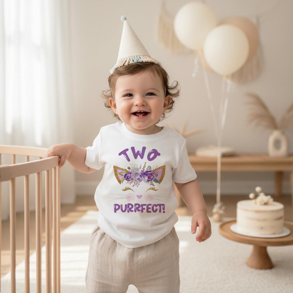 May include: A white short-sleeved t-shirt featuring a purple and gold graphic of cat ears adorned with purple flowers, closed eyes with long lashes, and the text "TWO PURRFECT!" in purple lettering, worn with a beige party hat.