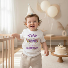 May include: A white short-sleeved t-shirt featuring a purple and gold graphic of cat ears adorned with purple flowers, closed eyes with long lashes, and the text "TWO PURRFECT!" in purple lettering, worn with a beige party hat.