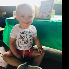 May include: A white short-sleeved bodysuit featuring the text "BRAYDEN is a WILD ONE" above a graphic of a lion, tiger, and elephant wearing colorful party hats, paired with red shorts and gray shoes, worn by a young child.
