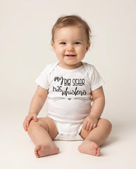 May include: A white short-sleeved baby bodysuit featuring the black printed text "my BIG SISTER has whiskers" with a small heart and thin horizontal lines resembling cat whiskers underneath the lettering on the front of the garment.