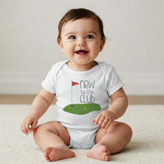 May include: A white short-sleeved baby bodysuit featuring a graphic of a green golf putting area with a red flag and the text "new to the CLUB" in dark gray lettering. The design is centered on the chest of the garment.
