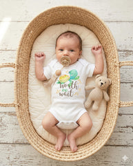 May include: A white baby bodysuit featuring a watercolor illustration of a green sea turtle and the text Worth the wait in brown and green script. The bodysuit is worn by a baby with a beige pacifier and a small crocheted tan teddy bear.