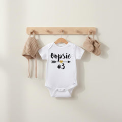 May include: A white short-sleeve baby bodysuit featuring the black text Oopsie, a gold glitter heart, an arrow graphic, and the number 3. The bodysuit is displayed on a wooden hanger alongside a matching tan knit bonnet and tan knit booties.